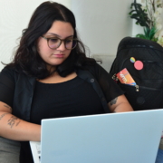 Mulher sentada em cadeira usando notebook. Ao fundo, uma bolsa preta e um vaso de flores.