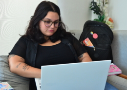 Mulher sentada em cadeira usando notebook. Ao fundo, uma bolsa preta e um vaso de flores.