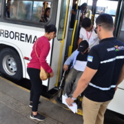 Uma mulher em pé, um homem cadeirante sendo ajudado por motoristas de ônibus para subir na plataforma de elevação, ao lado outro homem em pé.