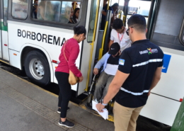 Uma mulher em pé, um homem cadeirante sendo ajudado por motoristas de ônibus para subir na plataforma de elevação, ao lado outro homem em pé.