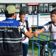 Homem entregando panfleto para dois alunos de escola estadual. Ao fundo, um ônibus na cor branca.