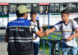 Homem entregando panfleto para dois alunos de escola estadual. Ao fundo, um ônibus na cor branca.