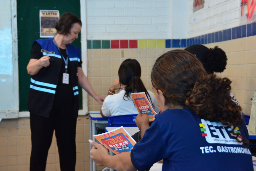 Alunas com panfleto na mão e ao fundo, mulher falando com outra aula. 