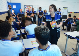 Mulher segurando panfleto e falando para alunos. Ao fundo, equipe de educação e fiscalização do Consórcio de Transporte Metropolitano