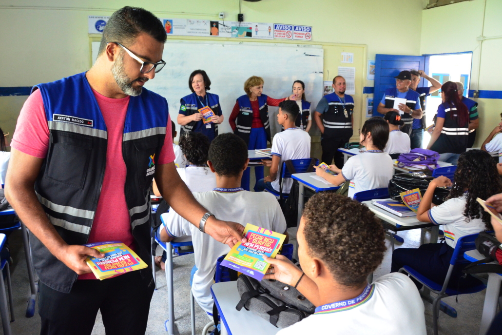 Homem fazendo panfletagem para alunos e alunas de escola estadual. Ao fundo equipe de educação e fiscalização do Consórcio de Transporte Metropolitano
