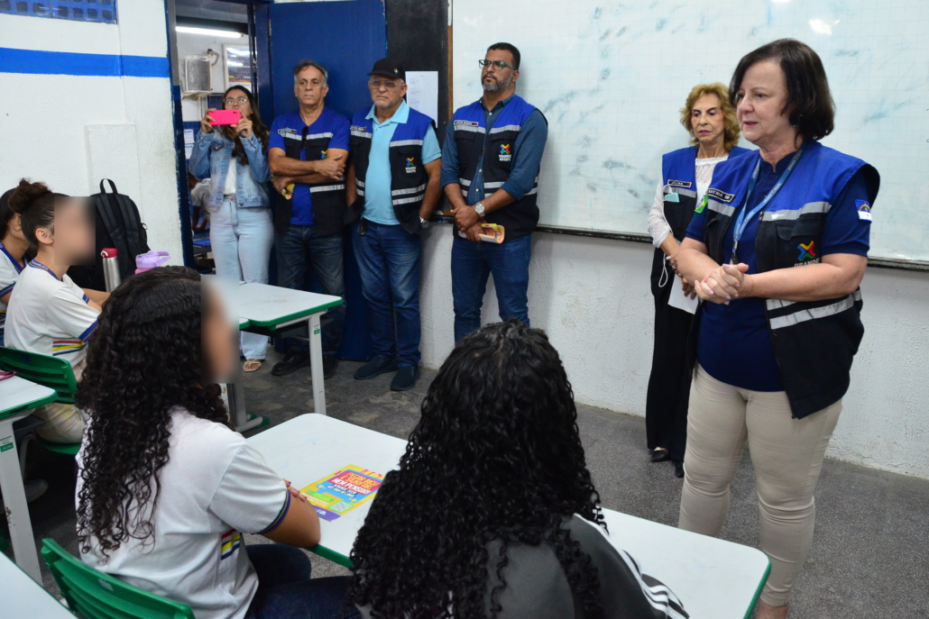 Mulher realizando palestras para quatro crianças do sexo feminino. Ao fundo, equipe escutando a palestra, são três homens e duas mulheres. O ambiente da foto é uma sala escolar. 