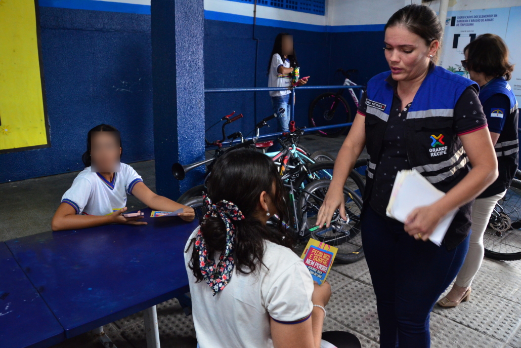 Mulher entregado panfleto para duas crianças do sexo feminino. Ao fundo, parede azul e branca. Bicicletas ao fundo. 