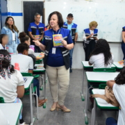 Mulher realizando palestras para crianças do sexo feminino e masculino. Ao fundo, equipe escutando a palestra, são dois homens e três mulheres. O ambiente da foto é uma sala escolar.