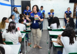 Mulher realizando palestras para crianças do sexo feminino e masculino. Ao fundo, equipe escutando a palestra, são dois homens e três mulheres. O ambiente da foto é uma sala escolar.