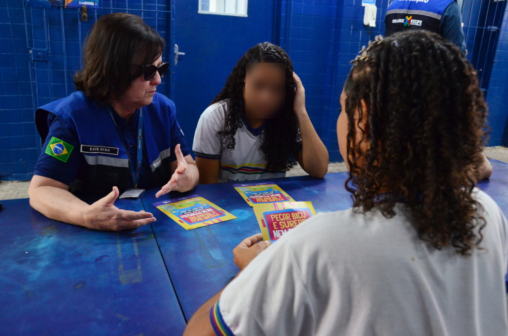Mulher falando com duas crianças do sexo feminino, elas estão sentadas, apoiadas em mesa na cor azul. Ao fundo, parede azul. 