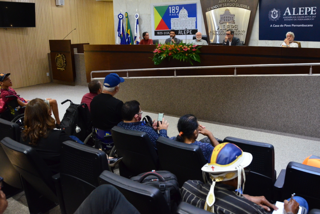 Auditório da Assembleia. Cinco pessoas sentadas na mesa plenária. Pessoas estão sentadas na plateia. 