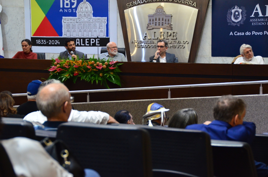 Auditório da Assembleia. Cinco pessoas sentadas na mesa plenária. Pessoas estão sentadas na plateia. 