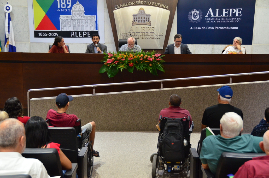 Auditório da Assembleia. Cinco pessoas sentadas na mesa plenária. Pessoas estão sentadas na plateia. 