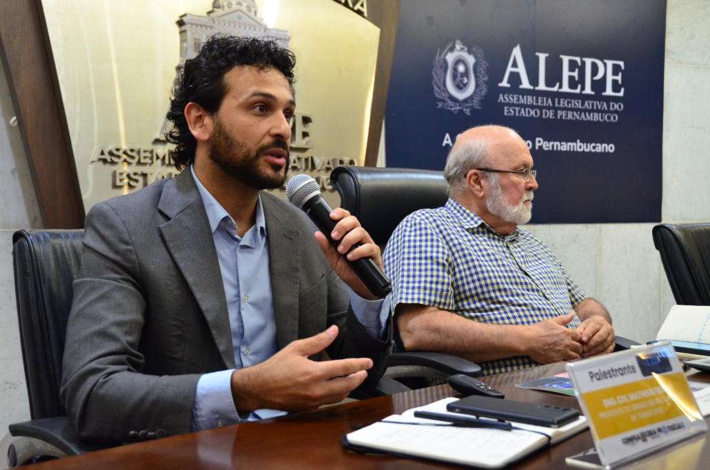 Perfil do Diretor-Presidente do Consórcio de Transporte Metropolitano, Matheus Freitas, que está falando no microfone. Ao seu lado, um homem idoso sentado. 