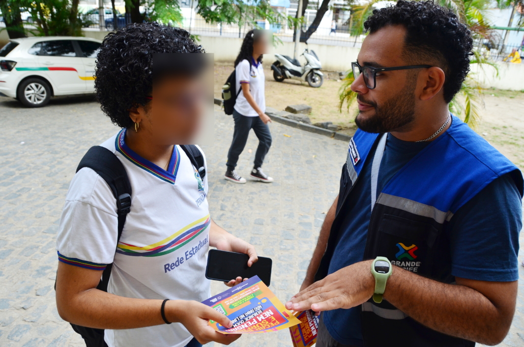 Homem entregando panfleto para criança do sexo feminino. O ambiente da foto é um pátio escolar. 