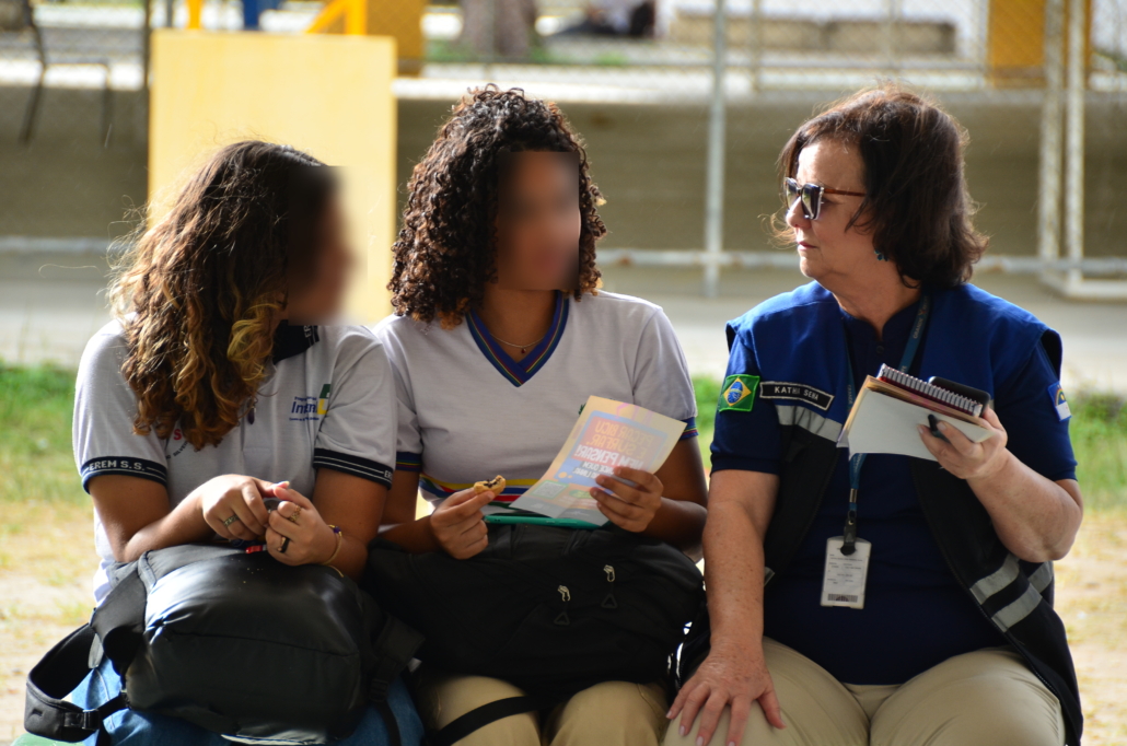Mulher entregado panfleto e conversando com duas crianças do sexo feminino. O ambiente da foto é um pátio escolar. 