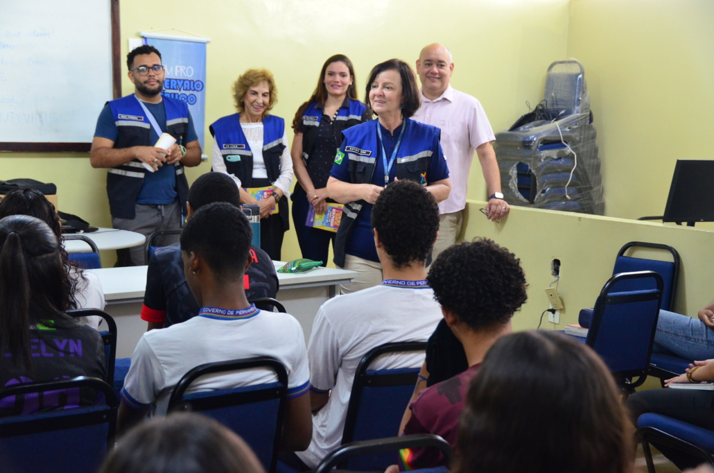 Mulher realizando palestra para crianças do sexo feminino e masculino. Ao fundo, equipe escutando a palestra, são dois homens e duas mulheres. O ambiente da foto é uma sala escolar.