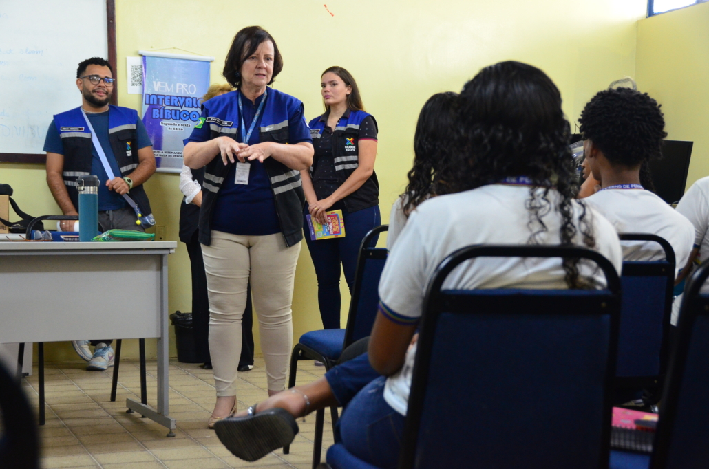 Mulher realizando palestra para crianças do sexo feminino e masculino. Ao fundo, equipe escutando a palestra, são dois homens e duas mulheres. O ambiente da foto é uma sala escolar.