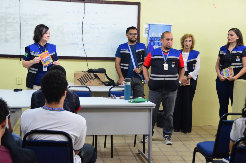 Homem realizando palestra para crianças do sexo feminino e masculino. Ao fundo, equipe escutando a palestra, são três mulheres e um homem. O ambiente da foto é uma sala escolar.