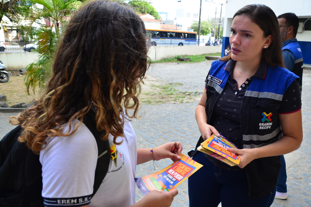 Mulher entregando panfleto para criança do sexo feminino. O ambiente da foto é um pátio escolar. 