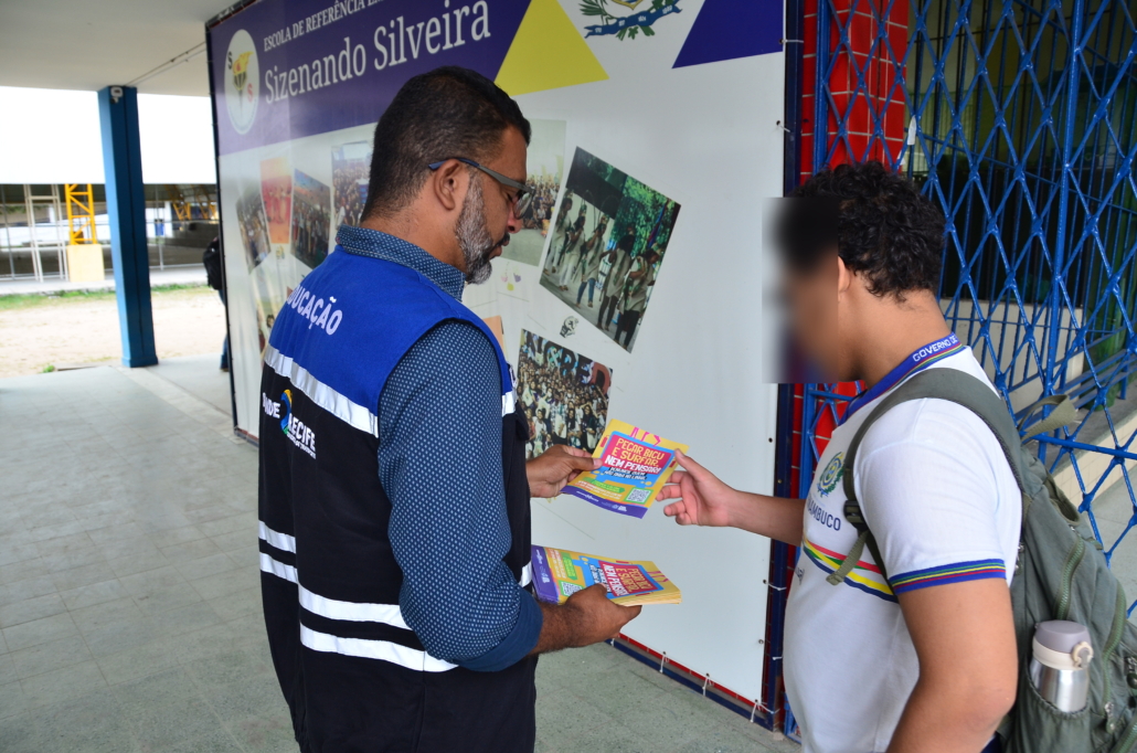 Homem entregando panfleto para criança do sexo masculino. O ambiente da foto é um pátio escolar. 