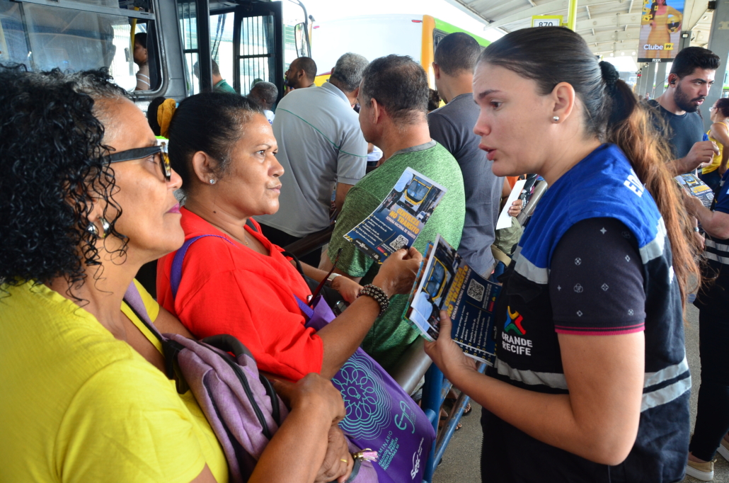 Mulher conversado e entregando panfleto para duas mulheres que estão na fila esperando ônibus