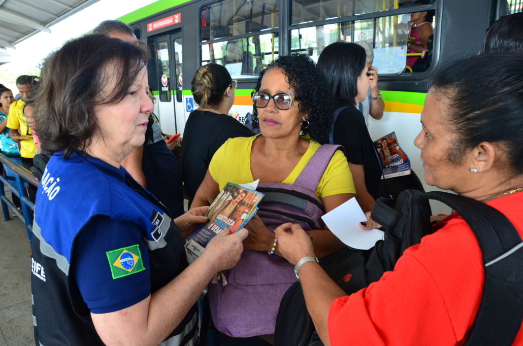 Mulher conversando e entregando panfleto para duas mulheres que estão na fila esperando ônibus